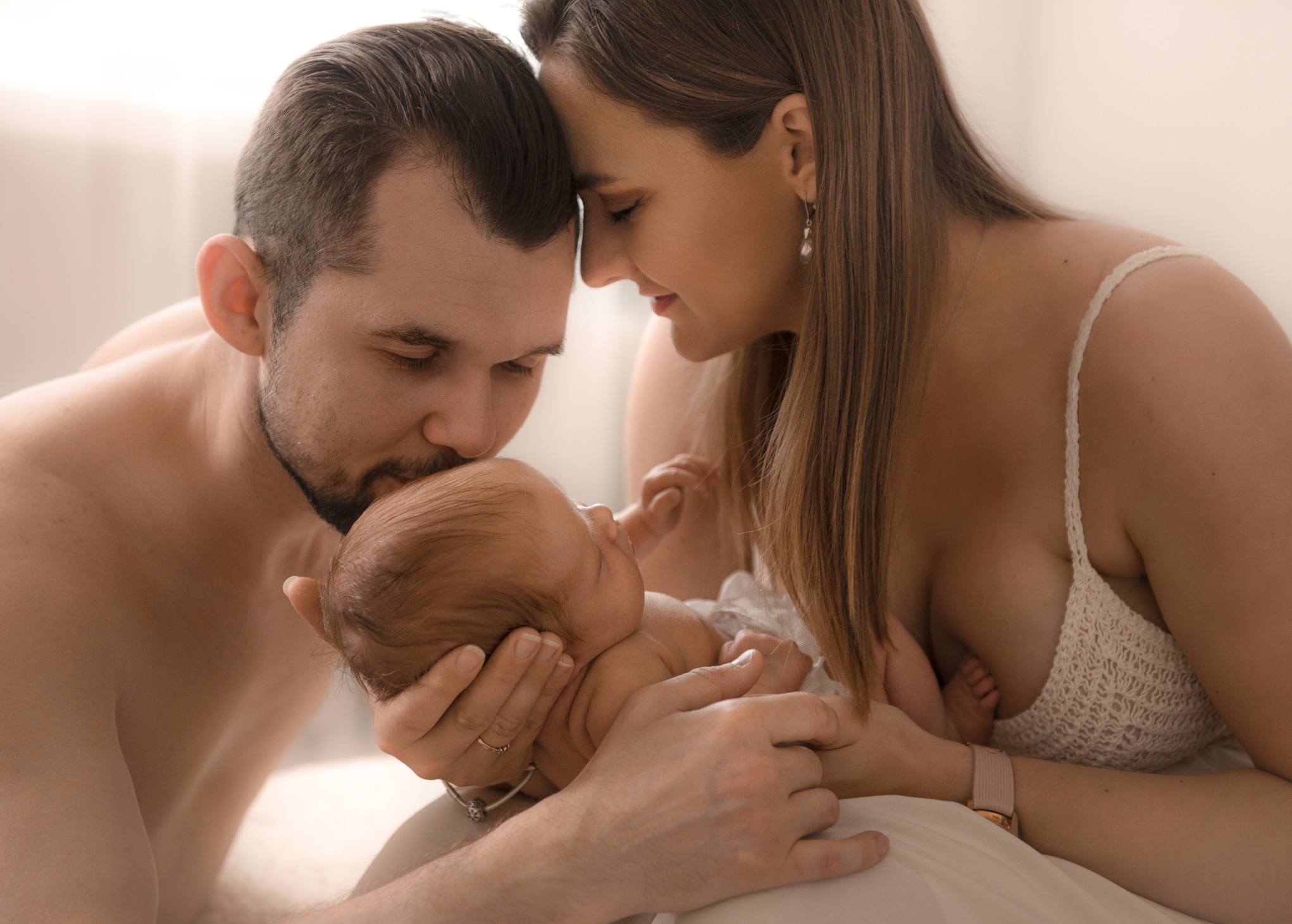 Rodzice tulący noworodka w czułym uścisku, delikatne światło podkreśla bliskość i intymność chwili, mama w koronkowej koszulce, tata z lekkim zarostem całuje główkę dziecka.