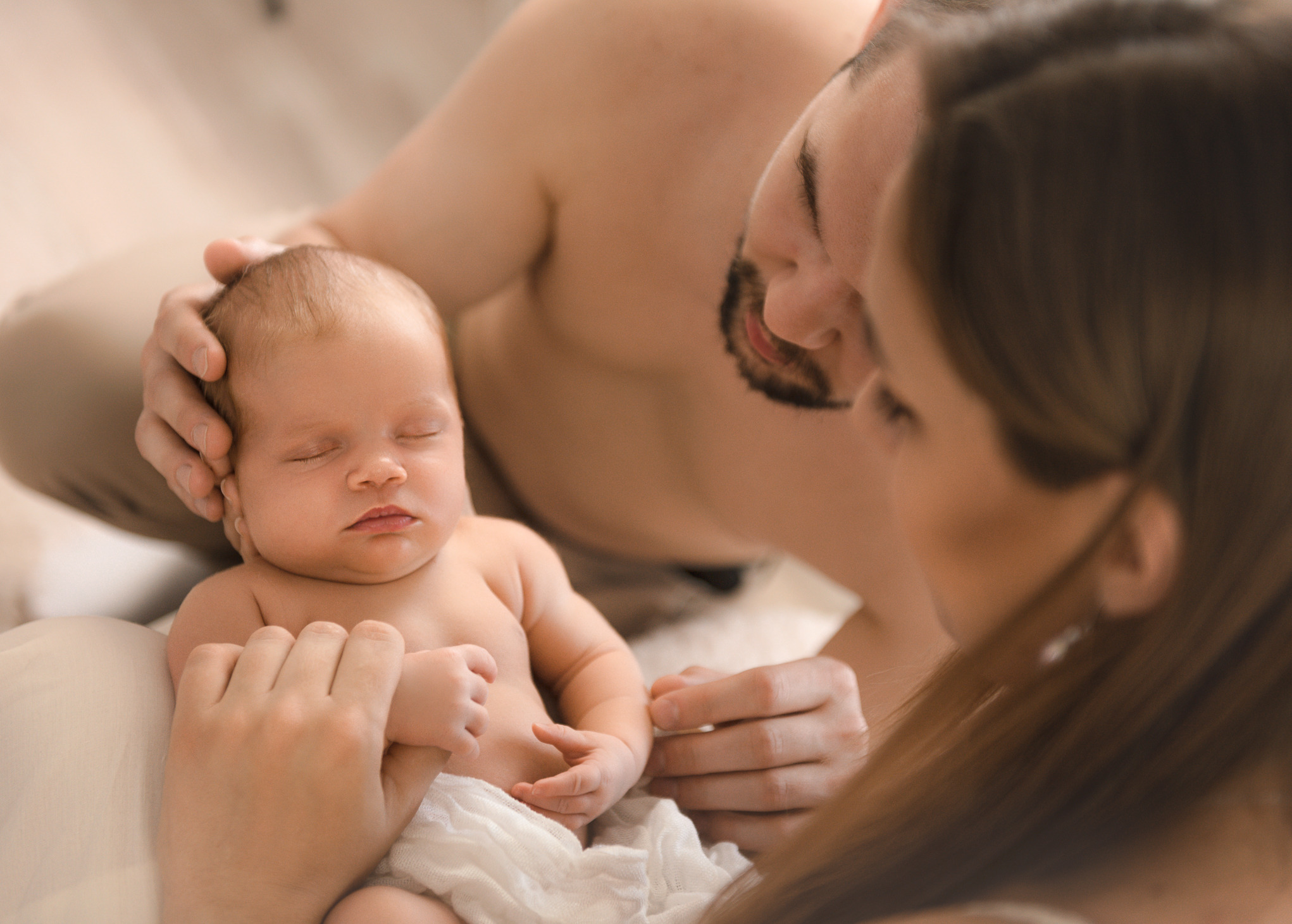 Śpiący noworodek w ramionach rodziców, delikatne oświetlenie, ciepłe barwy, tata z lekkim zarostem czule spogląda na dziecko.