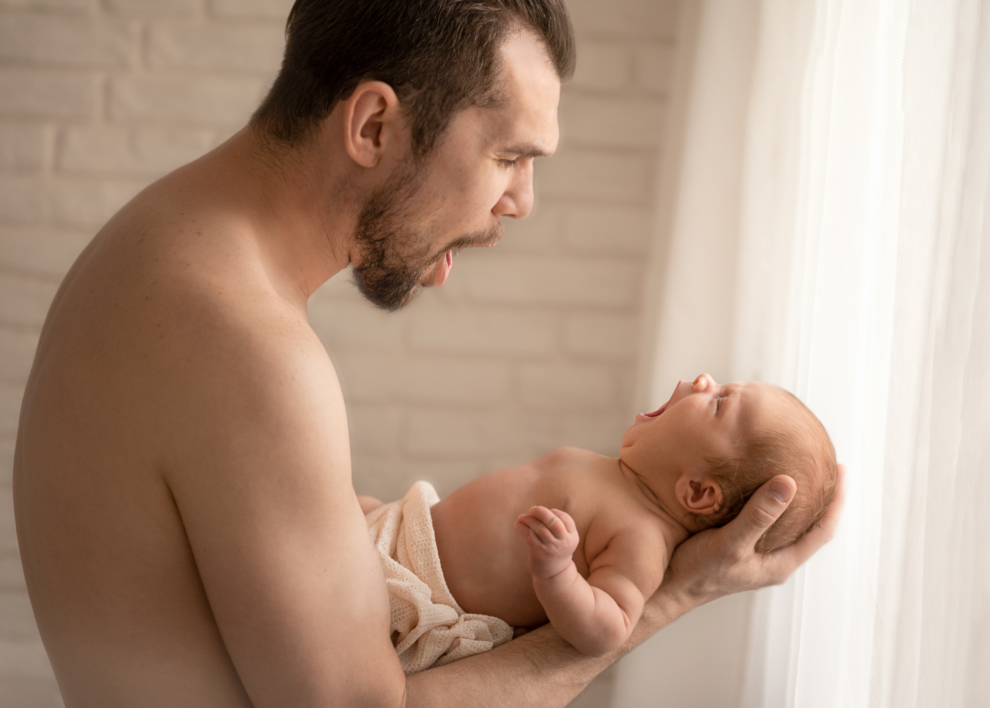Ojciec z brodą trzyma noworodka zawiniętego w beżowy kocyk, dziecko szeroko ziewa na tle jasnego okna i białej ceglanej ściany.