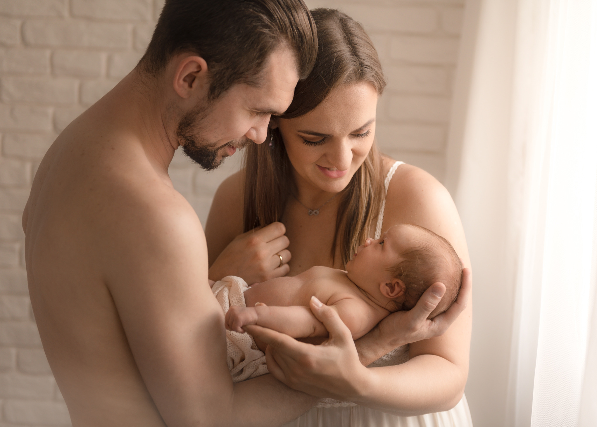 Szczęśliwi rodzice spoglądają z czułością na nowo narodzone dziecko, owinięte w kremowy kocyk, na tle jasnej ściany i okna.