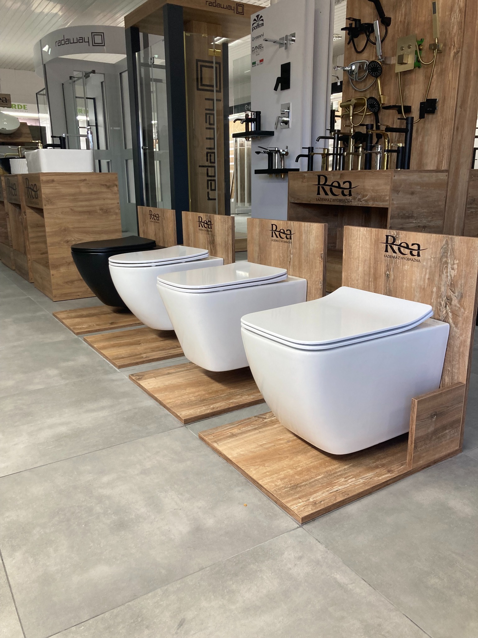 Ekspozycja czterech podwieszanych toalet w salonie łazienek, jedna czarna i trzy białe, każda z drewnianym panelem z logo Rea, na tle ekspozycji baterii i kabin prysznicowych.