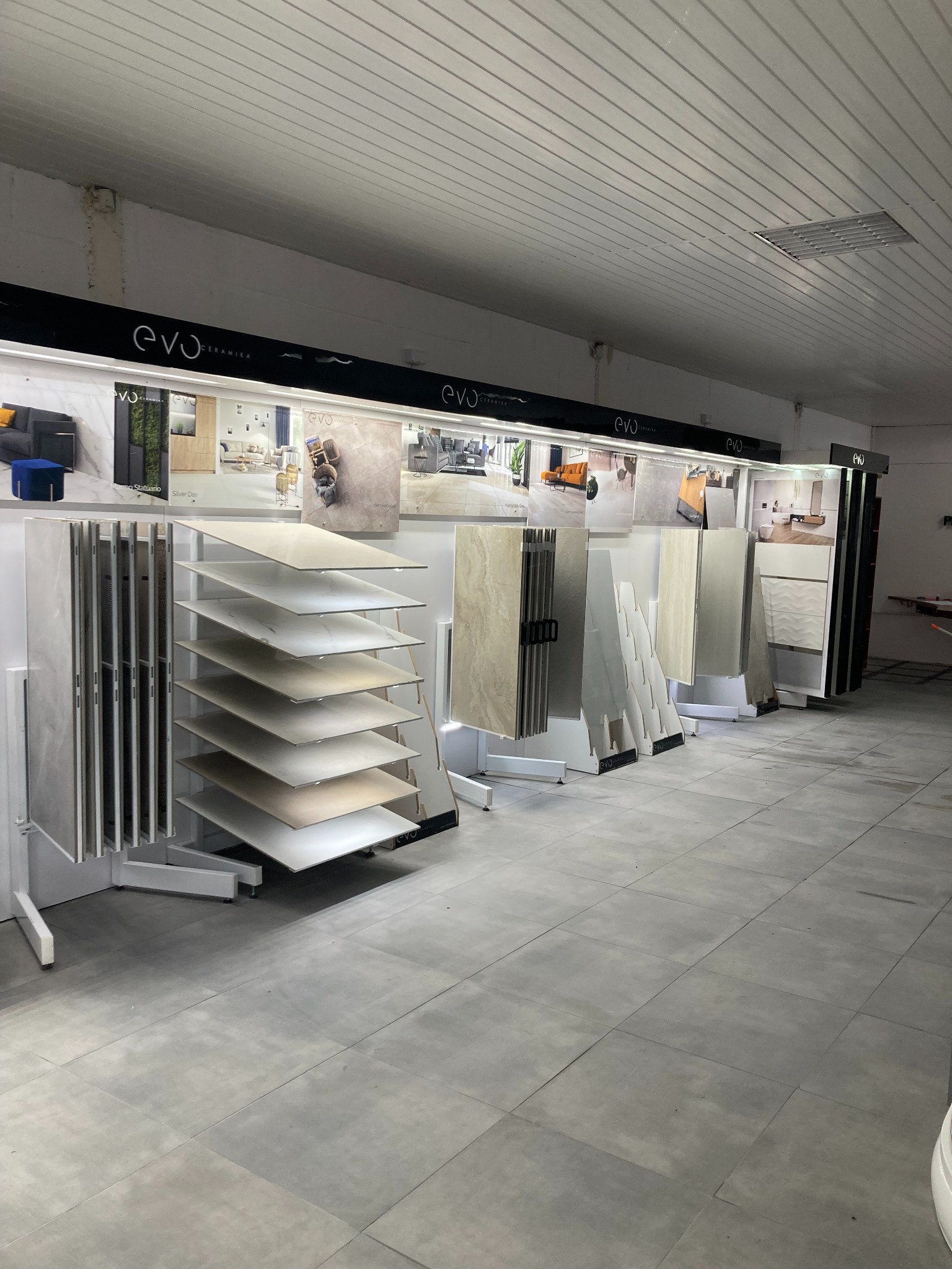 Ekspozycja płytek ceramicznych w salonie, różne wzory i kolory na stojakach i w formie aranżacji na górze, logo EVO Ceramika widoczne na górze ekspozycji.