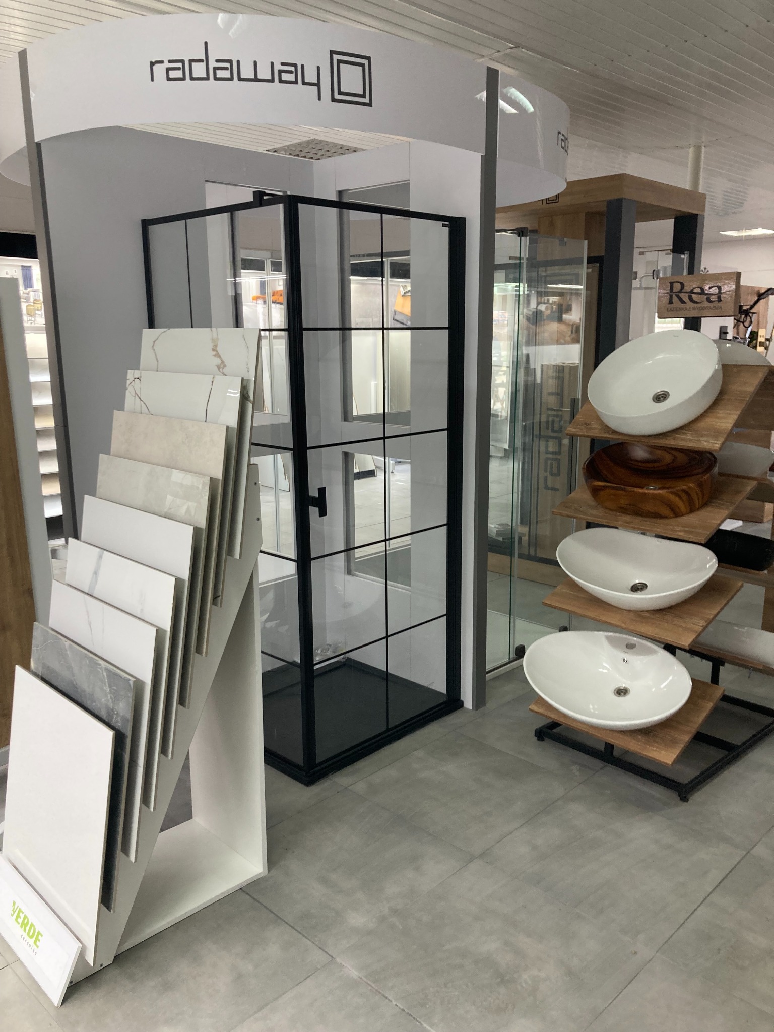 Ekspozycja w salonie łazienek: kabina prysznicowa z czarnymi profilami, próbki płytek ceramicznych na stojaku oraz umywalki nablatowe na drewnianych półkach.