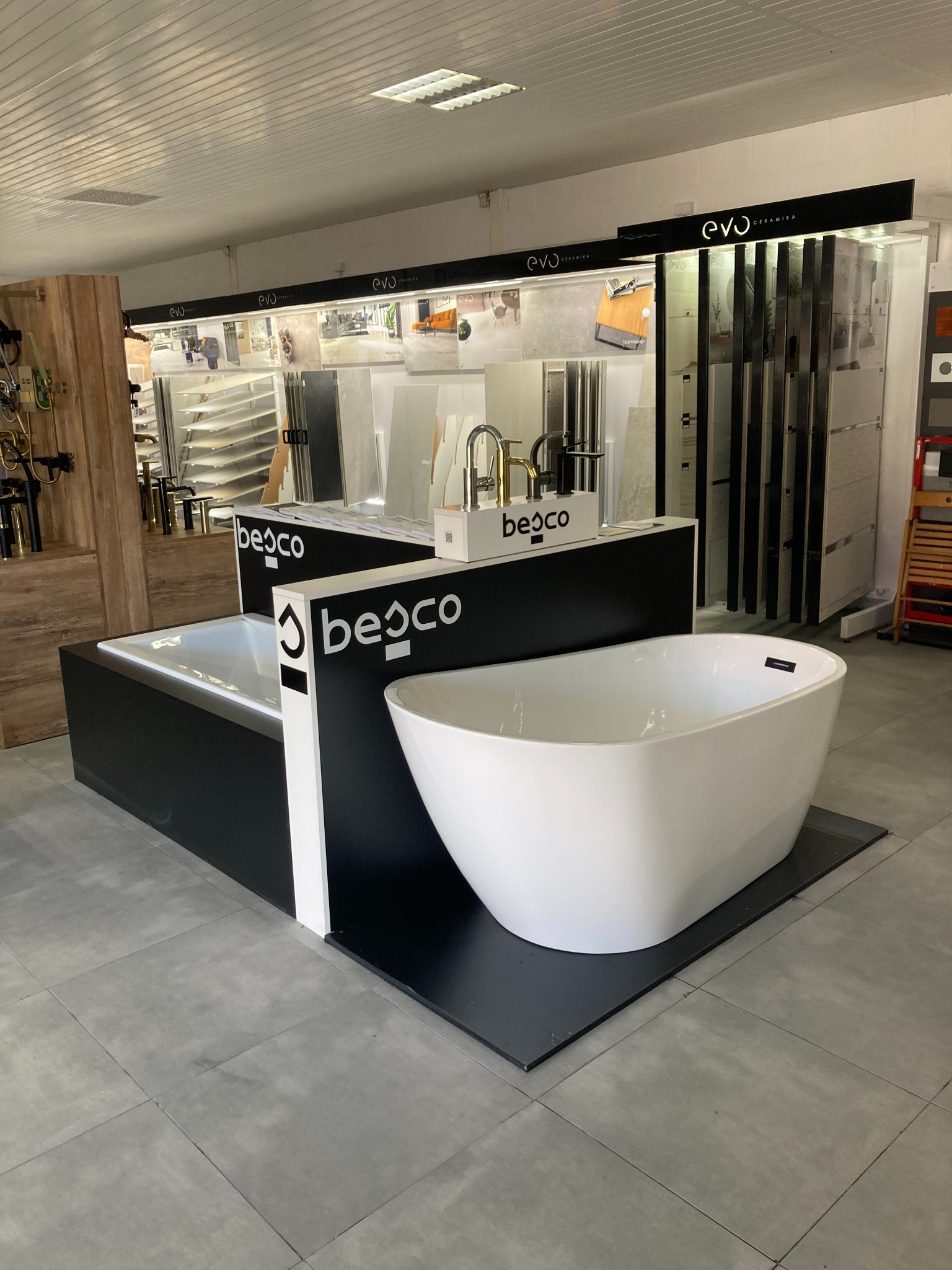 Ekspozycja w salonie łazienek: wolnostojąca wanna Besco o owalnym kształcie na czarnym podeście, umywalki nablatowe, baterie oraz płytki ceramiczne w tle.