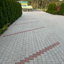 Ułożona kostka brukowa w odcieniach szarości z dekoracyjnym, zygzakowatym wzorem z czerwonej kostki, widok z perspektywy chodnika, w tle zieleń drzew i krzewów oraz zaparkowana ciężarówka.