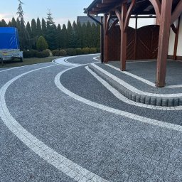 Mycie kostki brukowej - Ułożona kostka brukowa w odcieniach szarości z białymi pasami, tworząca wzór łuku. Widoczna drewniana wiata i zadbany ogród w tle. Precyzyjne wykonanie, estetyczny wygląd.