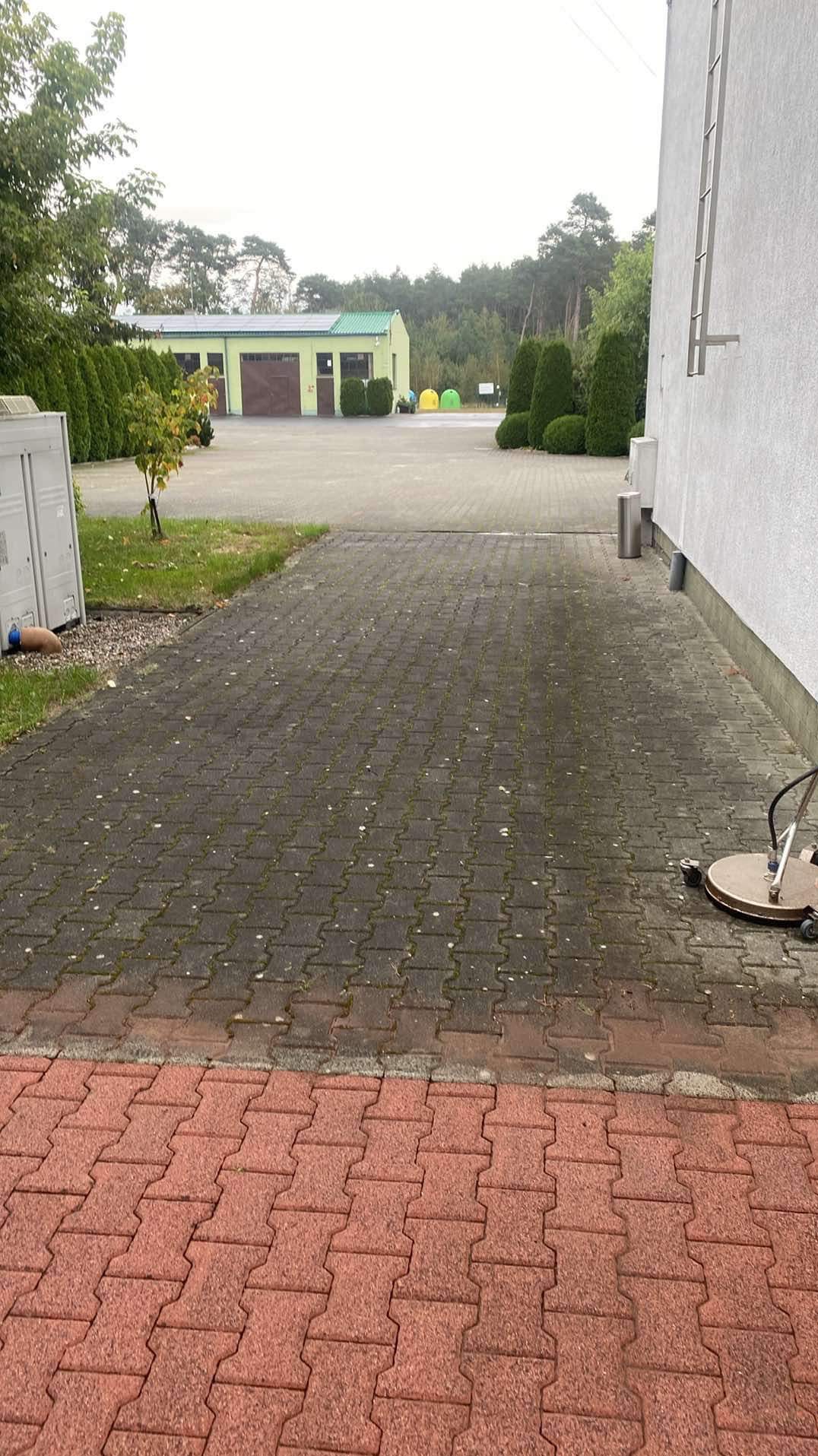 Częściowo wyczyszczona kostka brukowa przed budynkiem gospodarczym i ścianą, widoczna różnica w kolorze między czystą a brudną powierzchnią, w tle drzewa i krzewy.