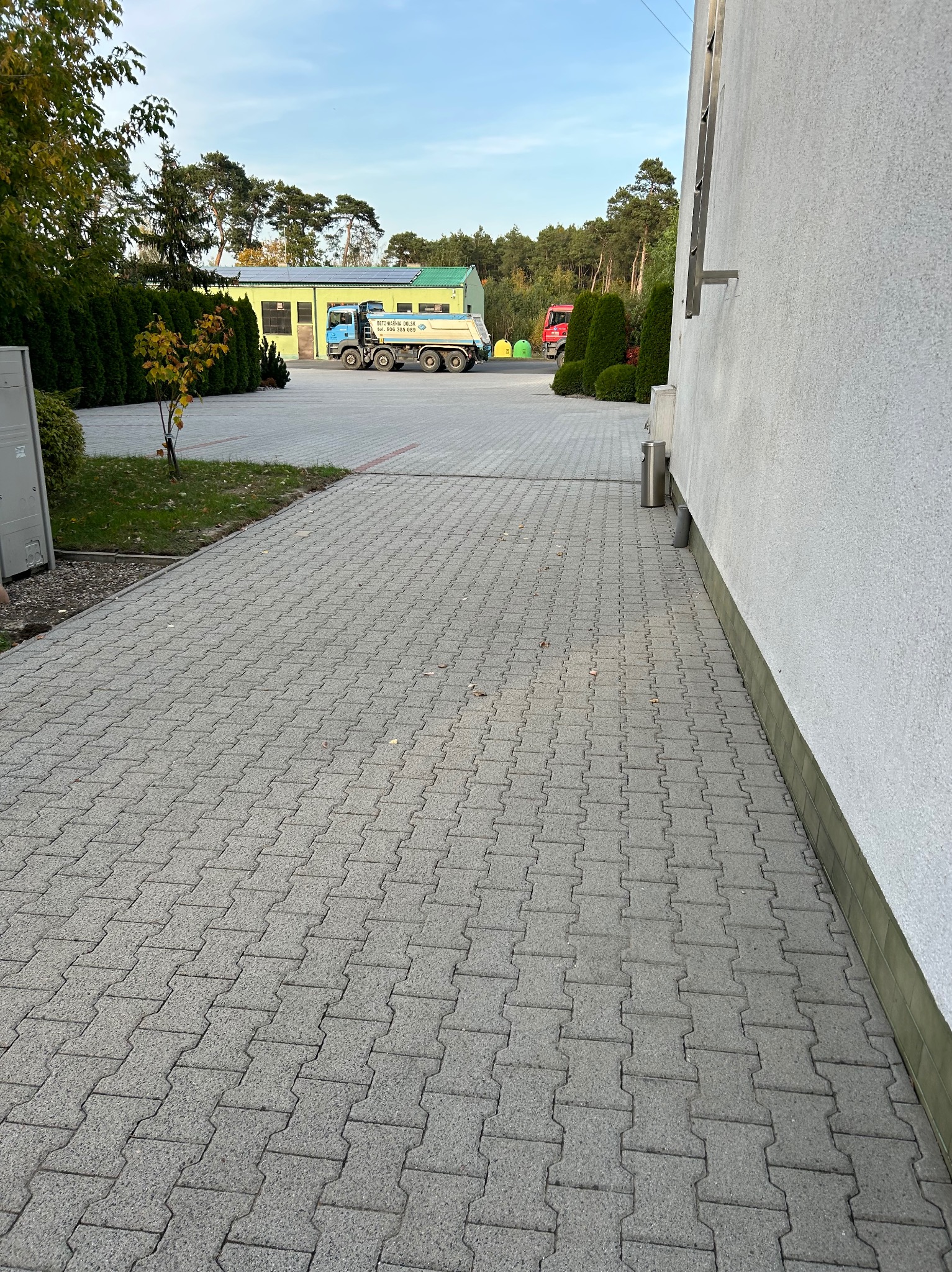 Szara kostka brukowa ułożona na parkingu przed budynkiem z widocznym samochodem ciężarowym w tle.