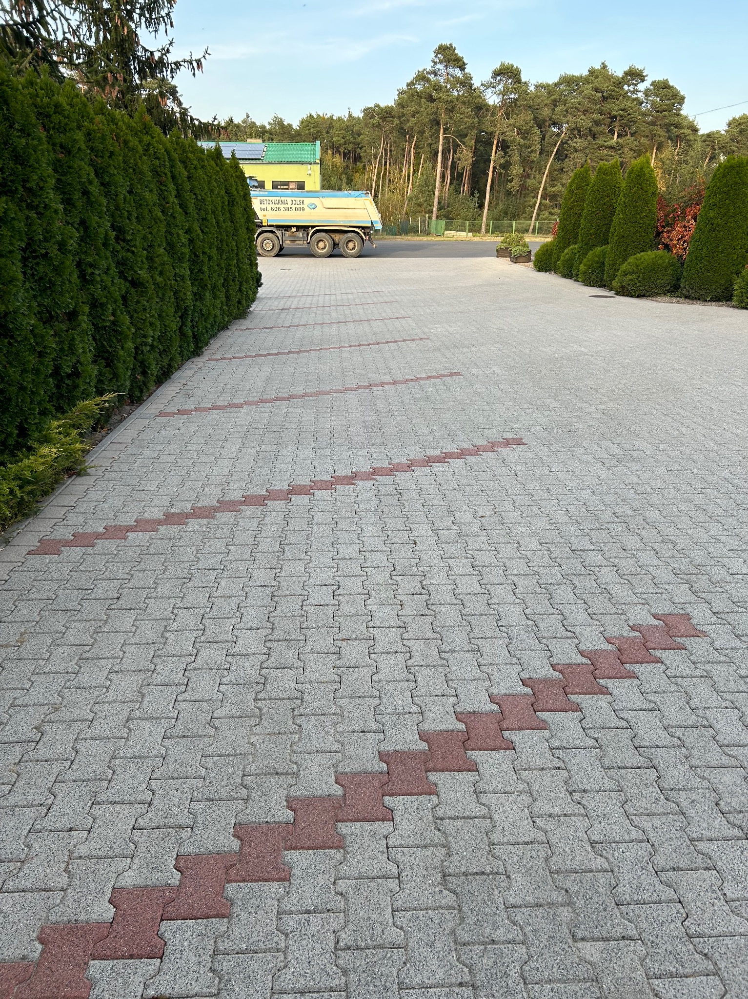 Ułożona kostka brukowa w odcieniach szarości z dekoracyjnym, zygzakowatym wzorem z czerwonej kostki, widok z perspektywy chodnika, w tle zieleń drzew i krzewów oraz zaparkowana ciężarówka.
