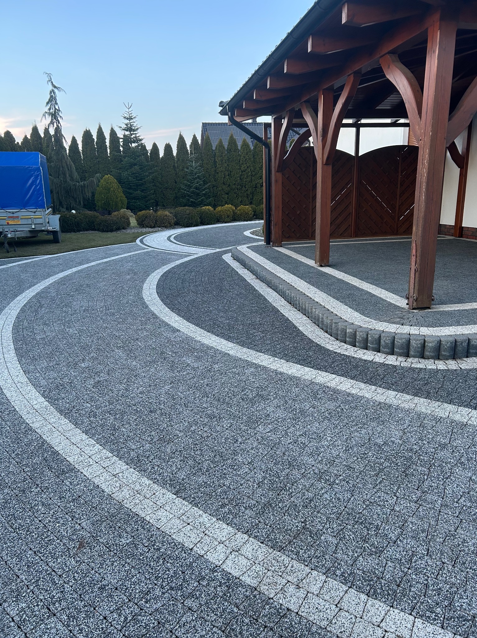 Ułożona kostka brukowa w odcieniach szarości z białymi pasami, tworząca wzór łuku. Widoczna drewniana wiata i zadbany ogród w tle. Precyzyjne wykonanie, estetyczny wygląd.