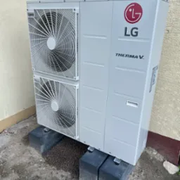 Zewnętrzna jednostka pompy ciepła LG Therma V, umieszczona na betonowych podstawach przy ścianie budynku o jasnej, teksturowanej elewacji. Widoczne rury instalacji grzewczej. Na ziemi rozsypane...