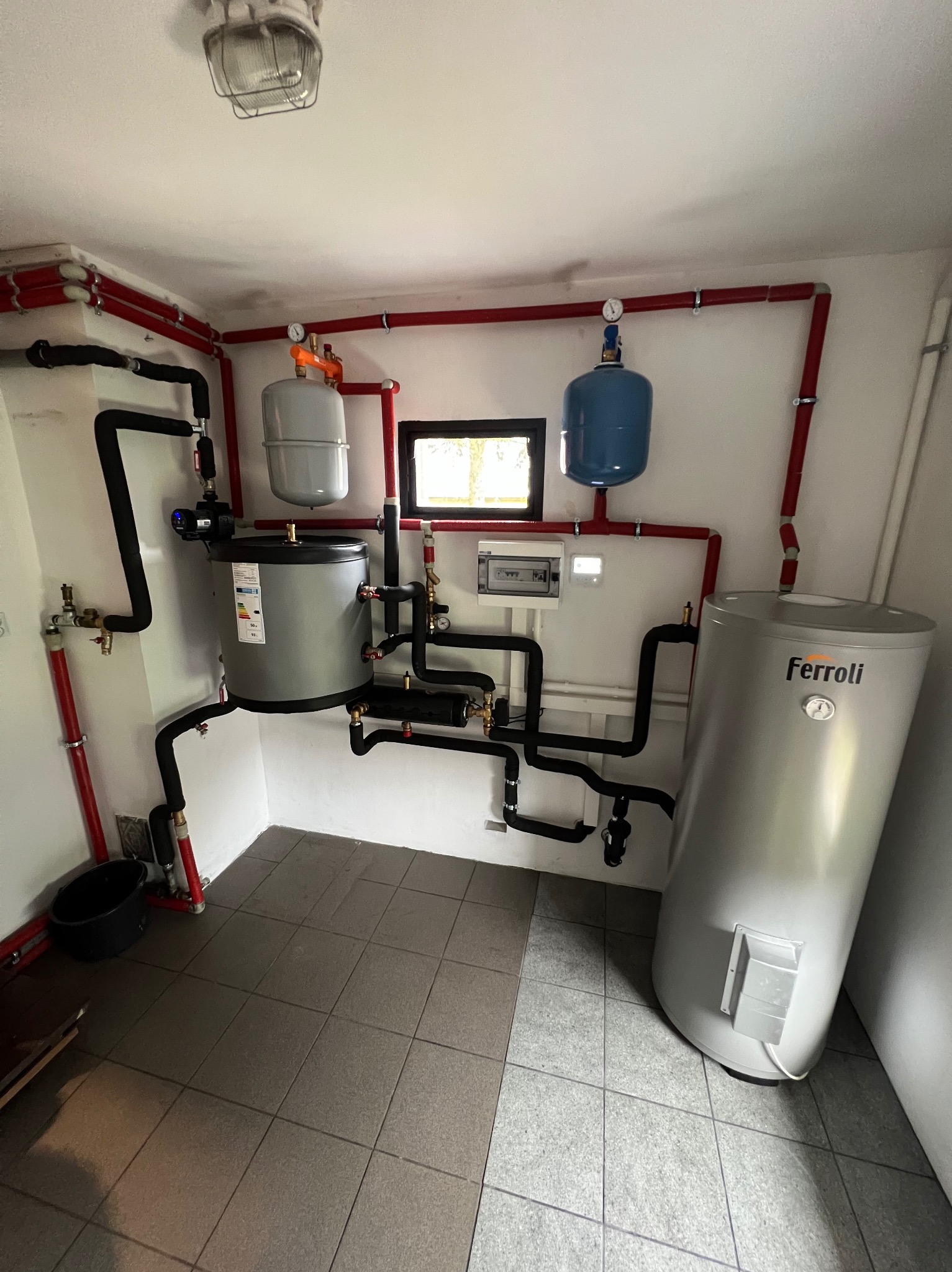 Kompletna instalacja kotłowni z dwoma zbiornikami, pompą obiegową i systemem rur w małym pomieszczeniu z szarą podłogą z płytek.