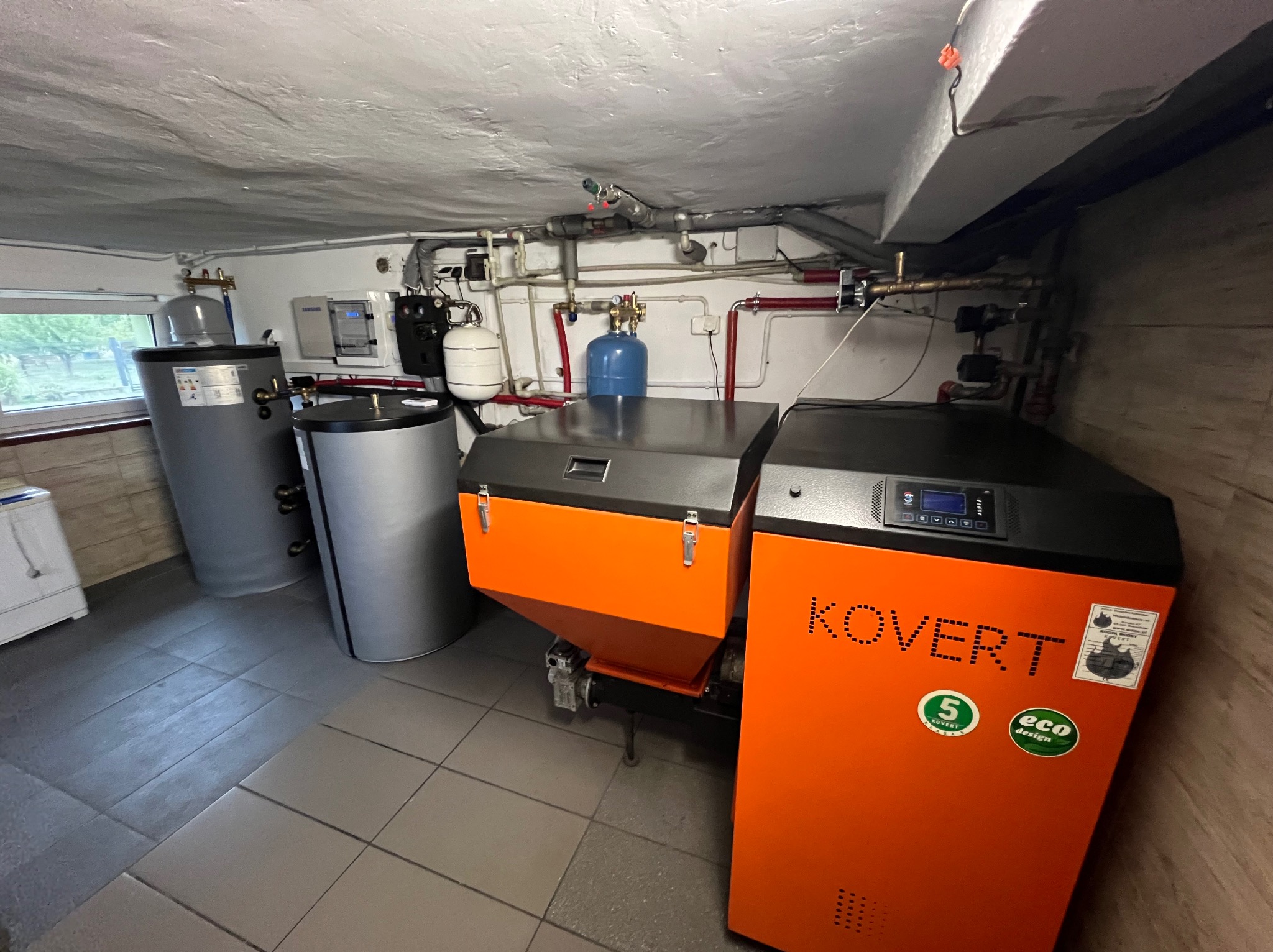 Pomieszczenie kotłowni z dwoma zbiornikami akumulacyjnymi i kotłem na paliwo stałe marki Kovert z podajnikiem, widoczne instalacje hydrauliczne i elektryczne.