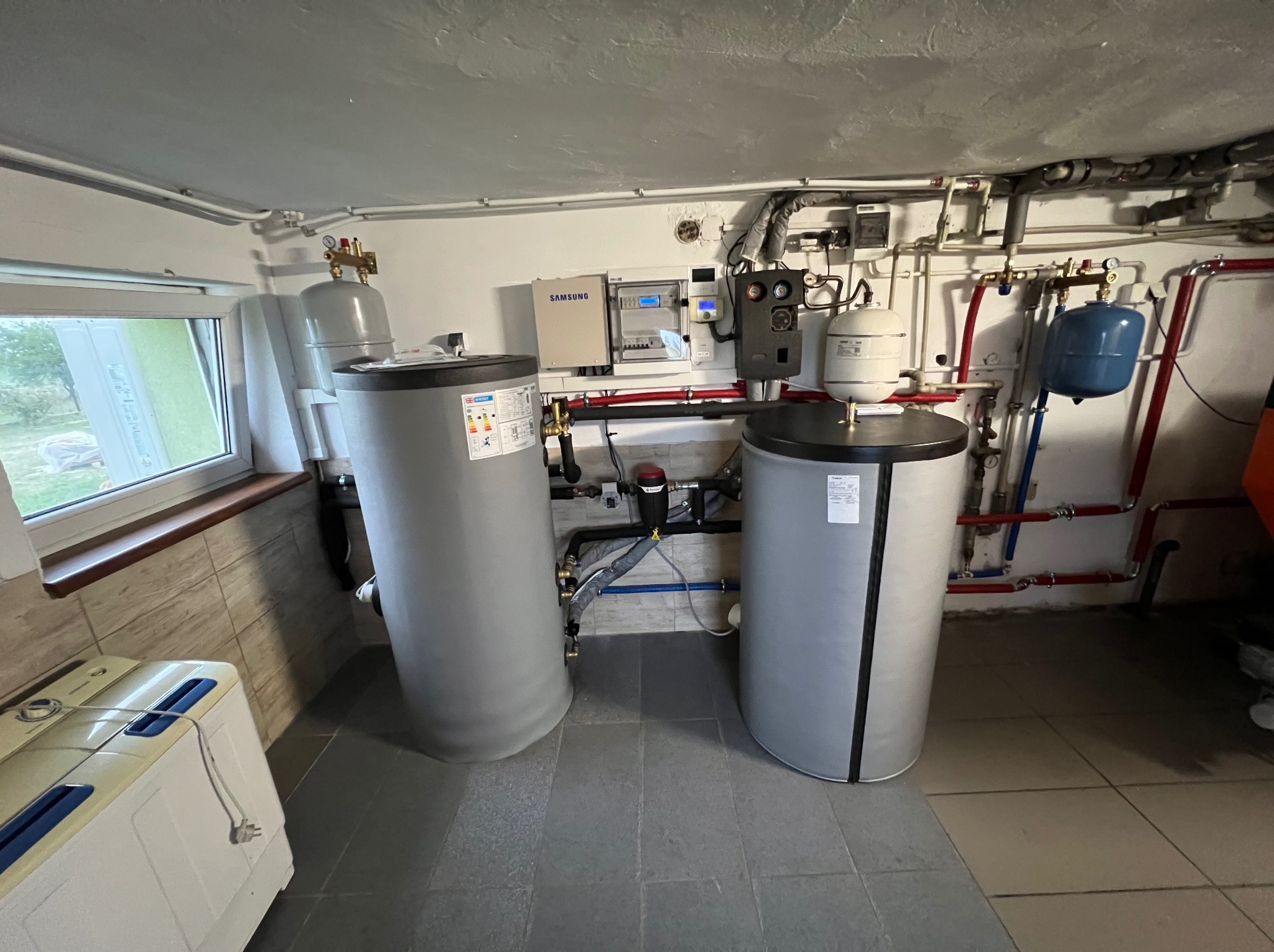 Dwa szare zbiorniki akumulacyjne ciepła w kotłowni z widoczną instalacją hydrauliczną i panelem sterowania Samsung na ścianie.
