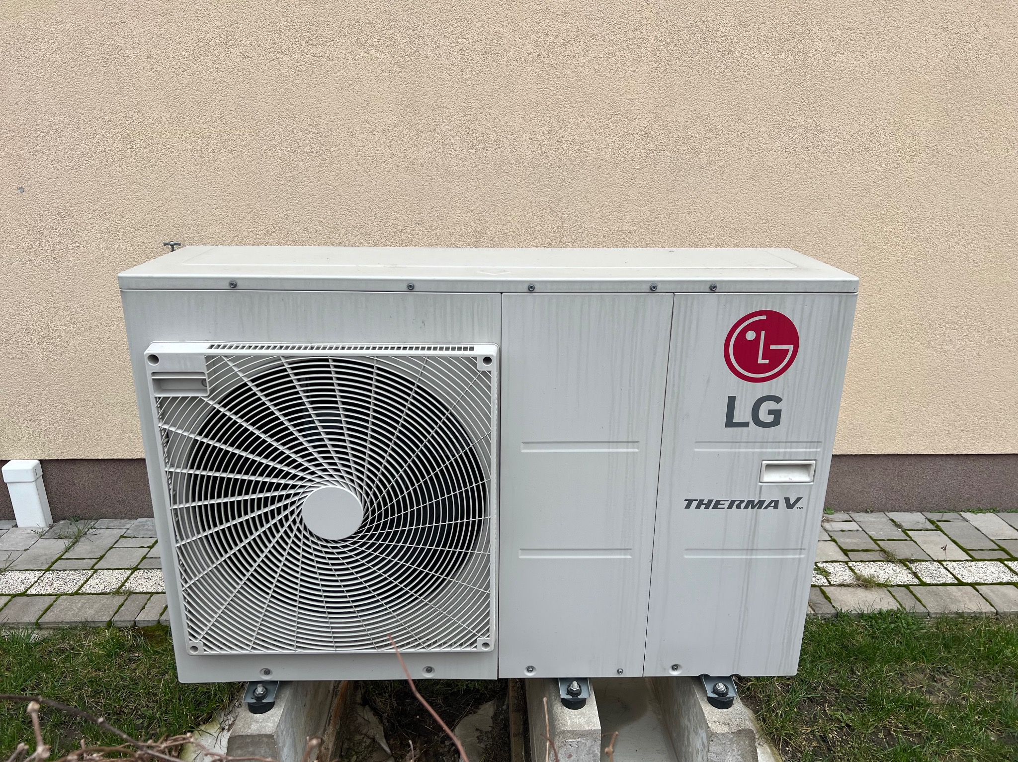 Zewnętrzna jednostka pompy ciepła LG Therma V, zamontowana na betonowym fundamencie przy ścianie budynku o beżowej elewacji. Widoczne ślady zabrudzeń na obudowie.