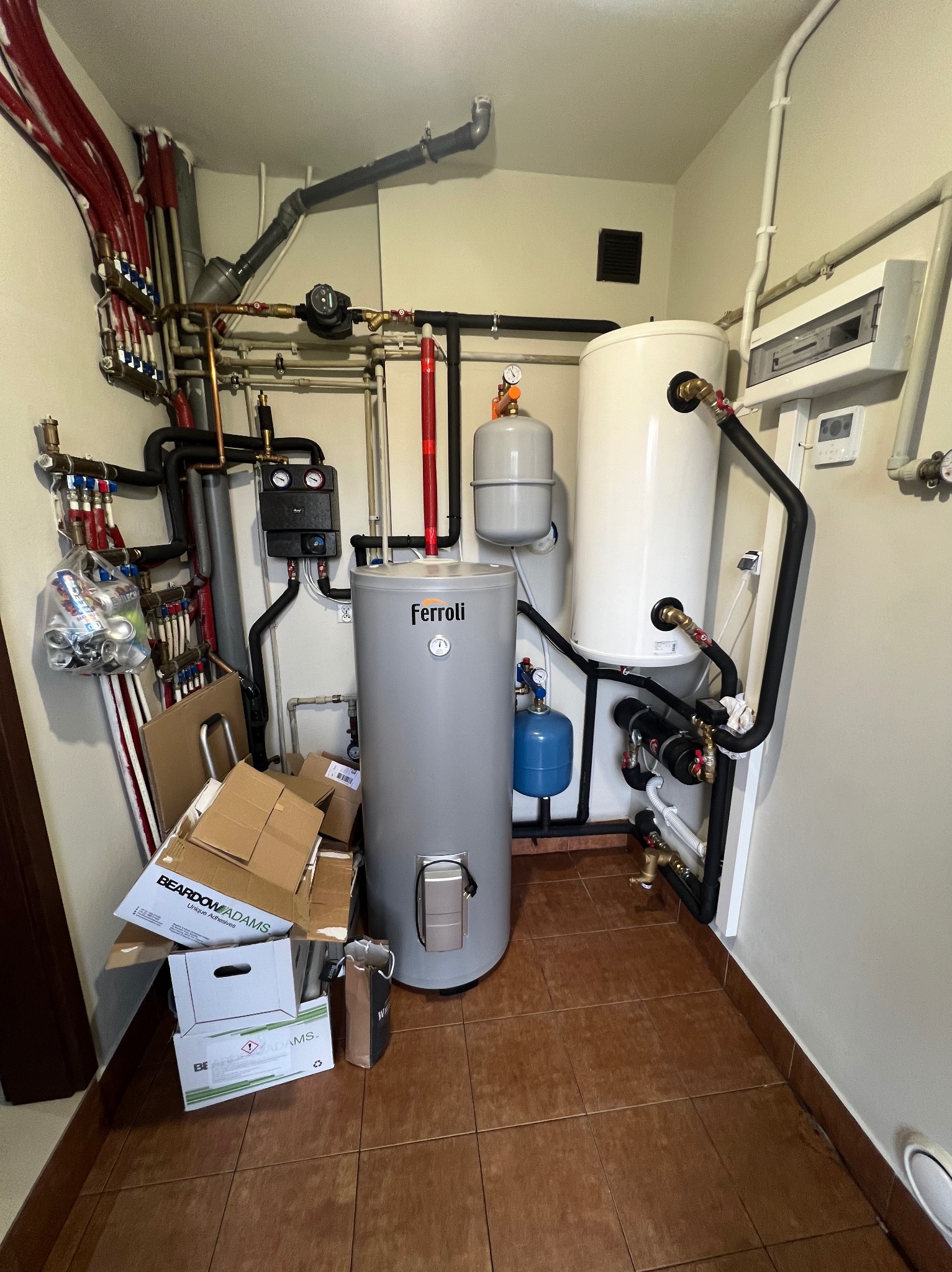 Kotłownia z szarym zasobnikiem Ferroli na pierwszym planie, biały zbiornik buforowy w tle, liczne rury miedziane i plastikowe, skrzynki kartonowe z napisem BEARDOW ADAMS na podłodze z terakoty.