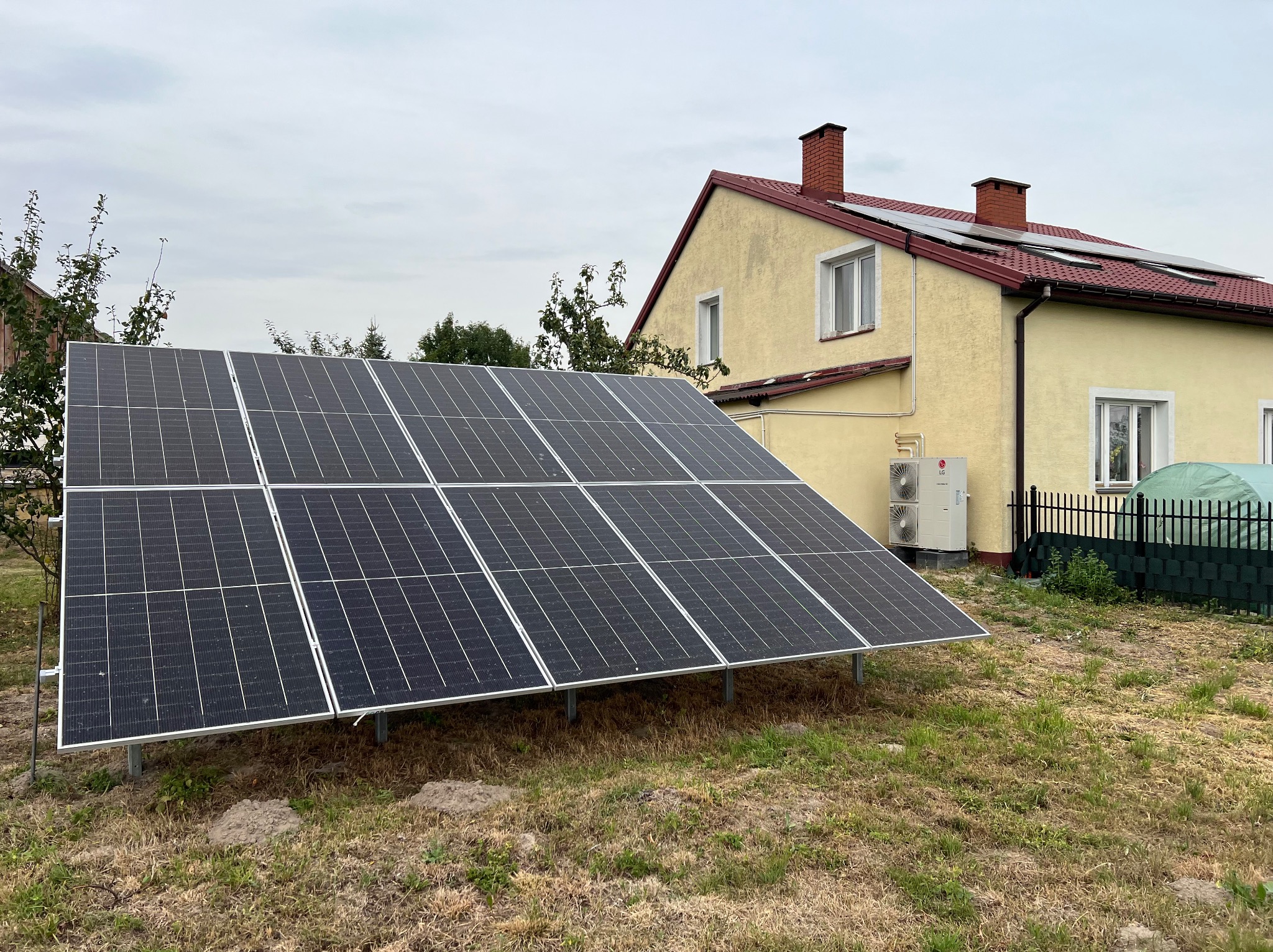 Instalacja fotowoltaiczna na gruncie przed domem z panelami na dachu, widoczne jednostki zewnętrzne klimatyzacji LG na ścianie budynku.