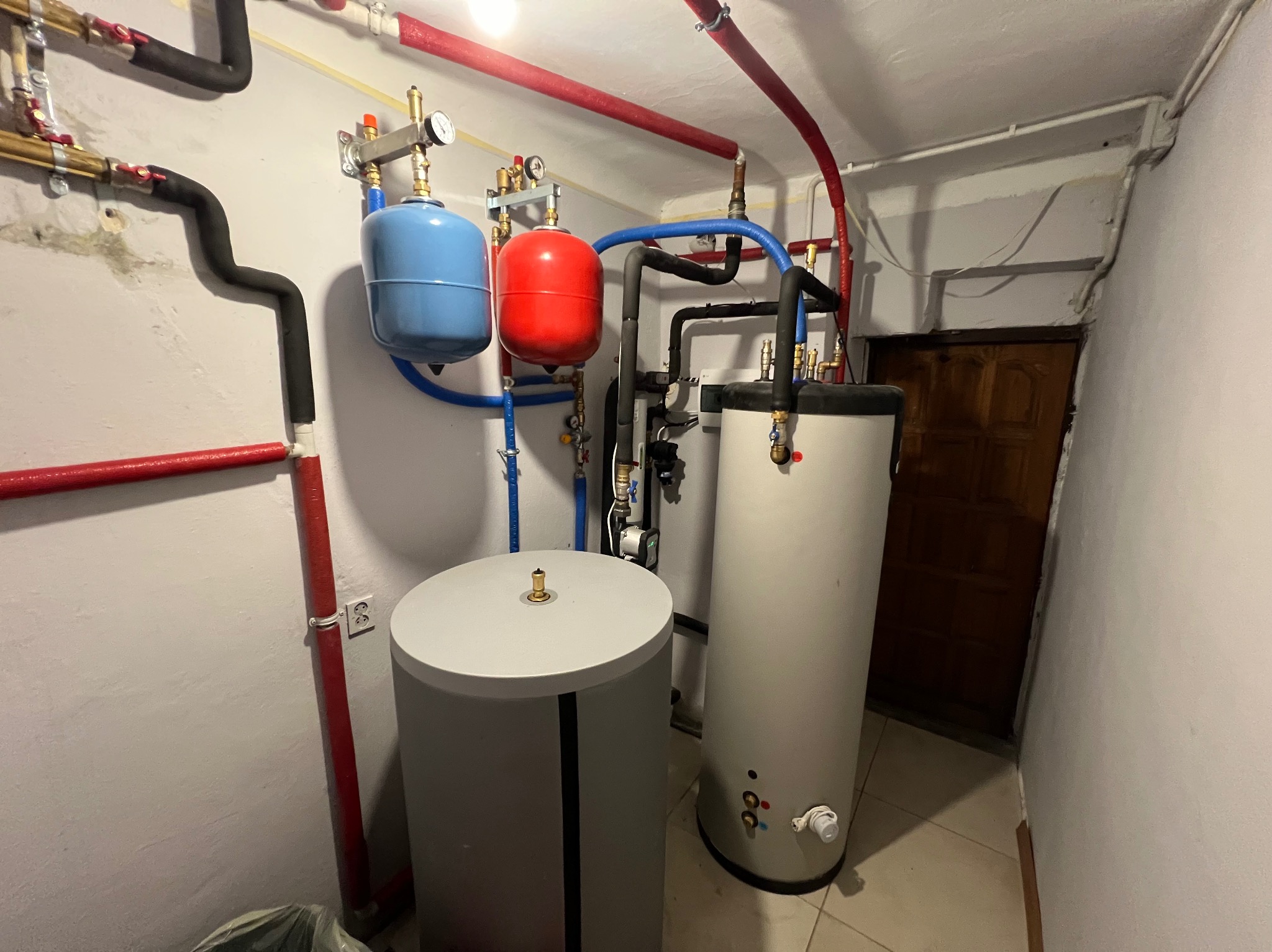 Instalacja pompy ciepła w kotłowni: dwa zbiorniki buforowe (szary i biały), czerwony i niebieski zbiornik ciśnieniowy, rury w izolacji czerwonej i czarnej, zawory i manometry.