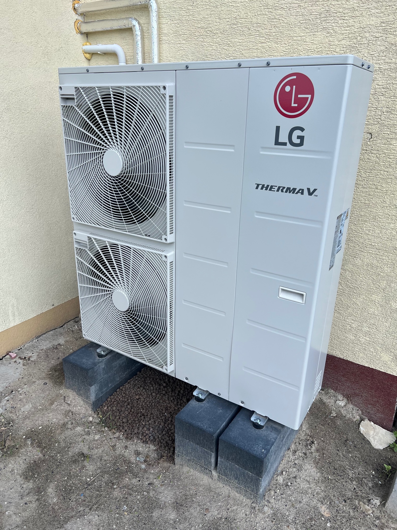 Zewnętrzna jednostka pompy ciepła LG Therma V, umieszczona na betonowych podstawach przy ścianie budynku o jasnej, teksturowanej elewacji. Widoczne rury instalacji grzewczej. Na ziemi rozsypane...