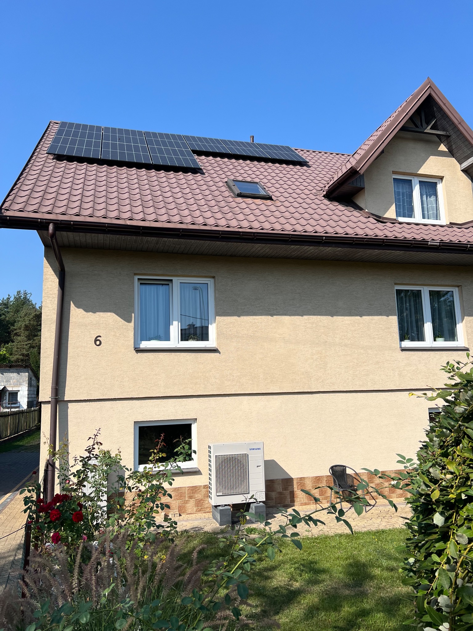 Dwukondygnacyjny dom z beżową elewacją i brązowym dachem, na którym zamontowane są panele fotowoltaiczne. Na ścianie widoczny numer domu '6', a obok okna zewnętrzna jednostka klimatyzacji marki...