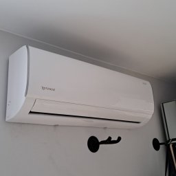 DARIUSZ KRÓL - Biała jednostka klimatyzacji marki Rotenso zamontowana na szarej ścianie nad czarnymi wieszakami na ubrania, widoczna również w odbiciu lustrzanym.