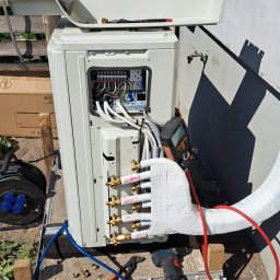 DARIUSZ KRÓL - Zewnętrzna jednostka klimatyzacji z otwartym panelem elektrycznym i podłączonymi rurami, częściowo owiniętymi izolacją. Widoczne narzędzia: wiertarko-wkrętarka akumulatorowa, kombinerki, manometr...