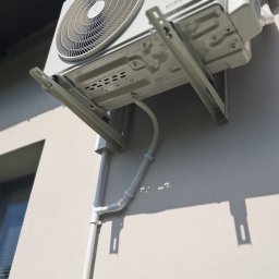 DARIUSZ KRÓL - Biała jednostka zewnętrzna klimatyzacji Rotenso, zamontowana na ścianie budynku w Konstancin-Jeziorna, widoczna z dołu pod kątem, z widocznymi rurami odprowadzającymi.