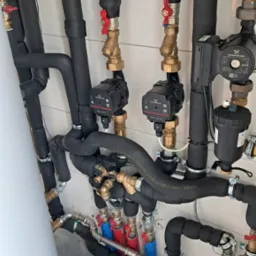 Instalacja grzewcza z dwoma pompami obiegowymi Grundfos Alpha L 25-40 180, rurami w izolacji termicznej, zaworami z czerwonymi pokrętłami i zbiornikiem akumulacyjnym.