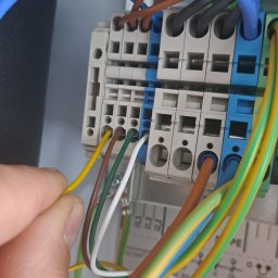 DARIUSZ KRÓL - Podłączenie przewodów elektrycznych do listwy zaciskowej wewnątrz skrzynki rozdzielczej, widoczne przewody w kolorach żółtym, brązowym, zielonym, białym i niebieskim, ręka instalatora wprowadza...