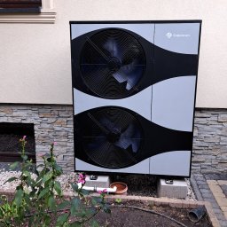DARIUSZ KRÓL - Zewnętrzna jednostka pompy ciepła Solplanet ustawiona na betonowych blokach przy ścianie domu z kamienną okładziną, obok rabata z roślinami i kamieniami.