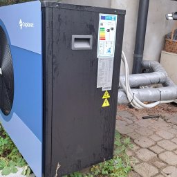 DARIUSZ KRÓL - Zewnętrzna jednostka pompy ciepła Solplanet, z widoczną etykietą energetyczną i naklejkami ostrzegawczymi, zainstalowana na kostce brukowej obok rur izolowanych taśmą, zarośnięta roślinnością.