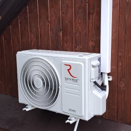 DARIUSZ KRÓL - Zewnętrzna jednostka klimatyzacji Rotenso Ukura zamontowana na drewnianej ścianie budynku z widocznymi elementami instalacji odprowadzającej skropliny i osłoną przewodów.