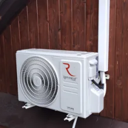 Zewnętrzna jednostka klimatyzacji Rotenso Ukura zamontowana na drewnianej ścianie budynku z widocznymi elementami instalacji odprowadzającej skropliny i osłoną przewod&oacute;w.