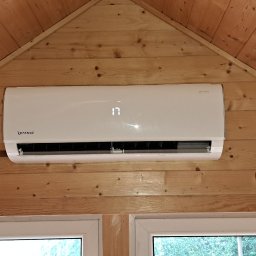 DARIUSZ KRÓL - Biały klimatyzator marki Rotenso zamontowany na drewnianej ścianie w kształcie trójkąta, widoczne okno z zielenią na zewnątrz.