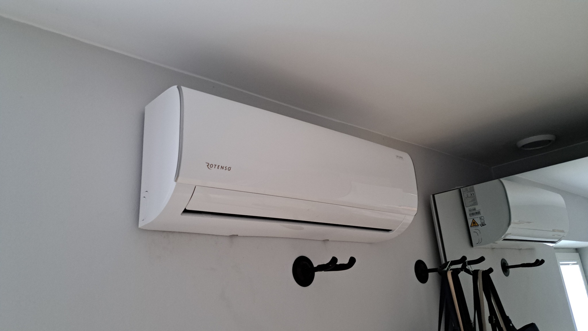 Biała jednostka klimatyzacji marki Rotenso zamontowana na szarej ścianie nad czarnymi wieszakami na ubrania, widoczna również w odbiciu lustrzanym.