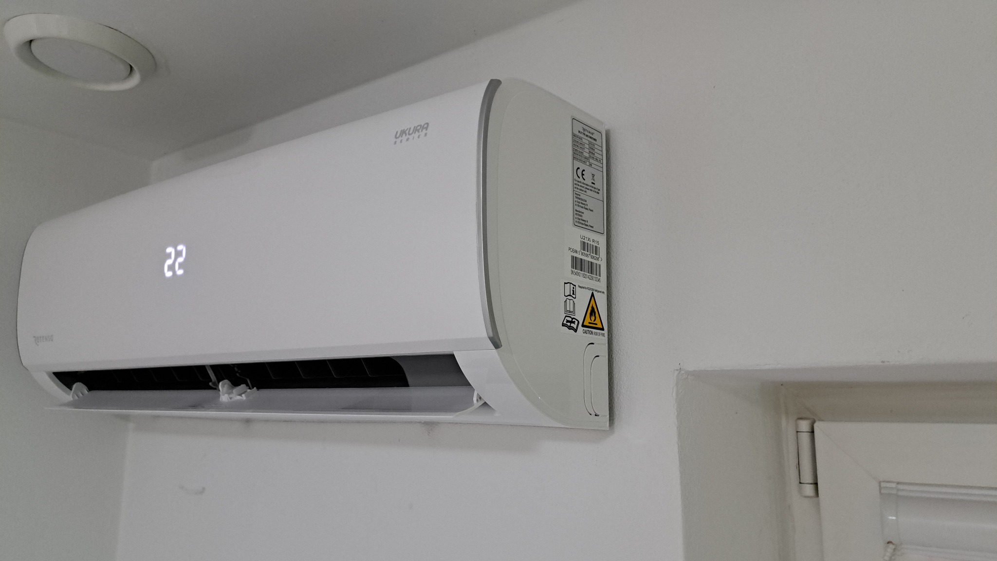 Biała klimatyzacja Ukura Series z wyświetlaczem temperatury 22 stopnie Celsjusza, zamontowana na białej ścianie w pomieszczeniu z widocznym fragmentem okna.