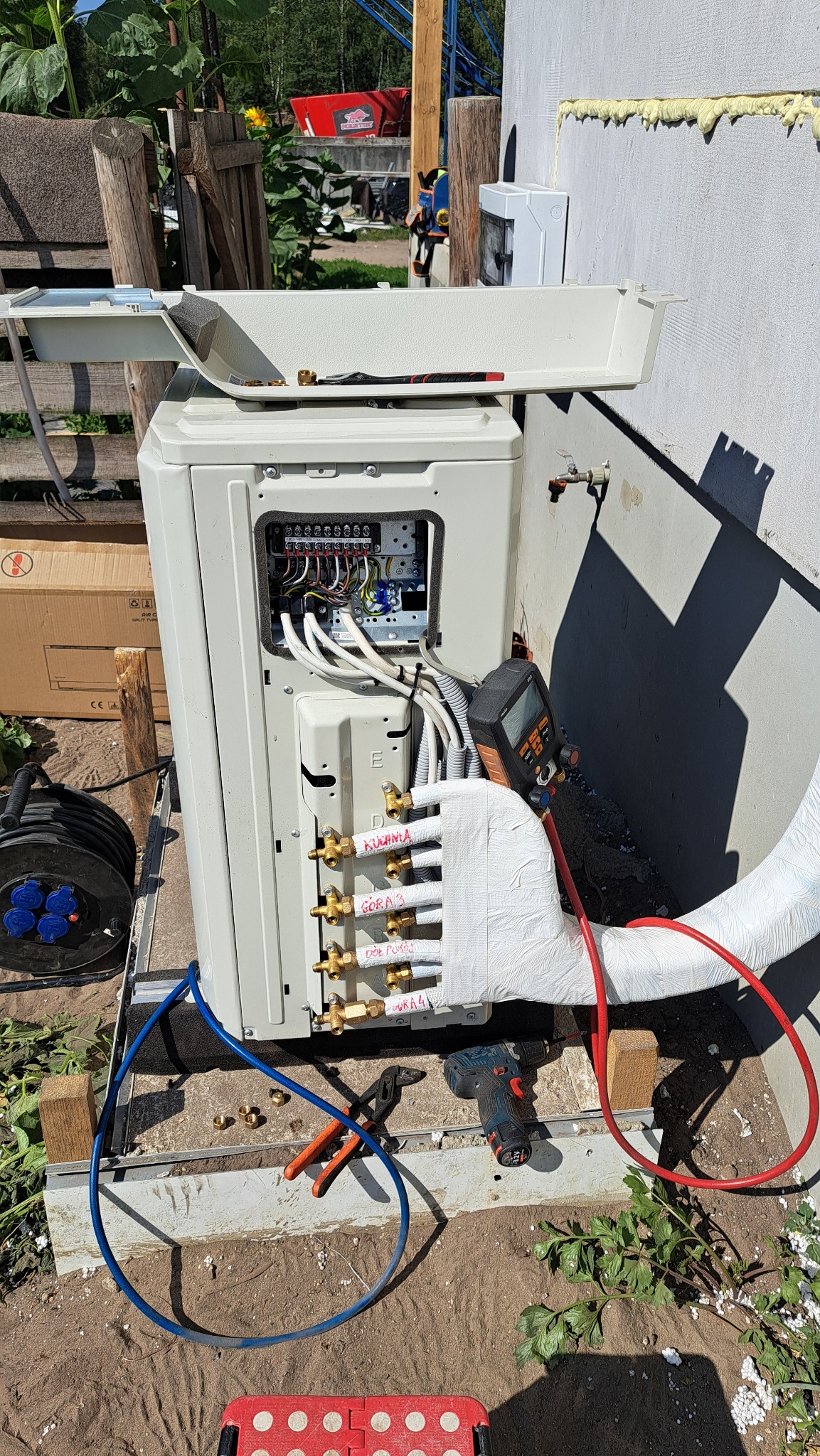 Zewnętrzna jednostka klimatyzacji z otwartym panelem elektrycznym i podłączonymi rurami, częściowo owiniętymi izolacją. Widoczne narzędzia: wiertarko-wkrętarka akumulatorowa, kombinerki, manometr...