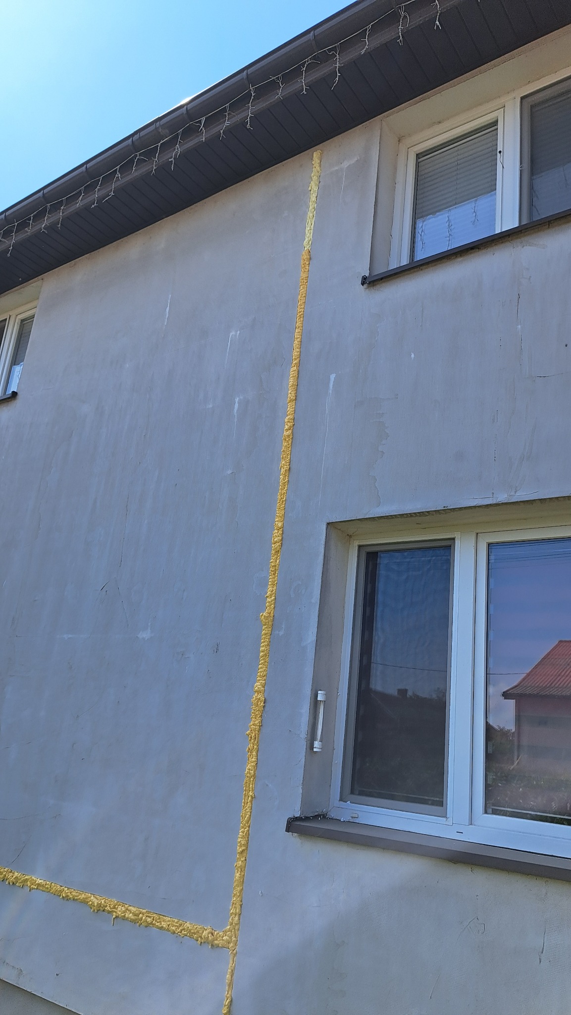 Ocieplanie ściany budynku z użyciem pianki montażowej wokół okien, widoczne ślady po pianie na szarej elewacji, okna z białymi ramami.