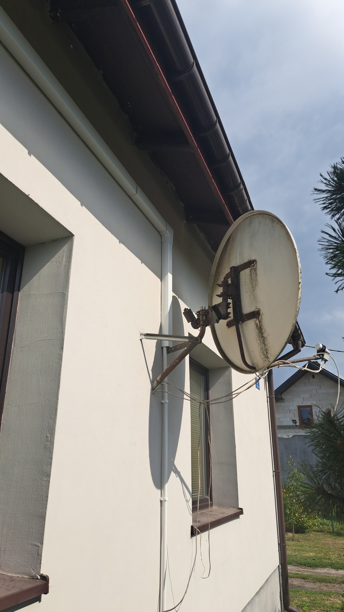 Biała antena satelitarna zamontowana na ścianie domu z widocznymi przewodami i elementami mocującymi, zarys okna i rynny w tle.