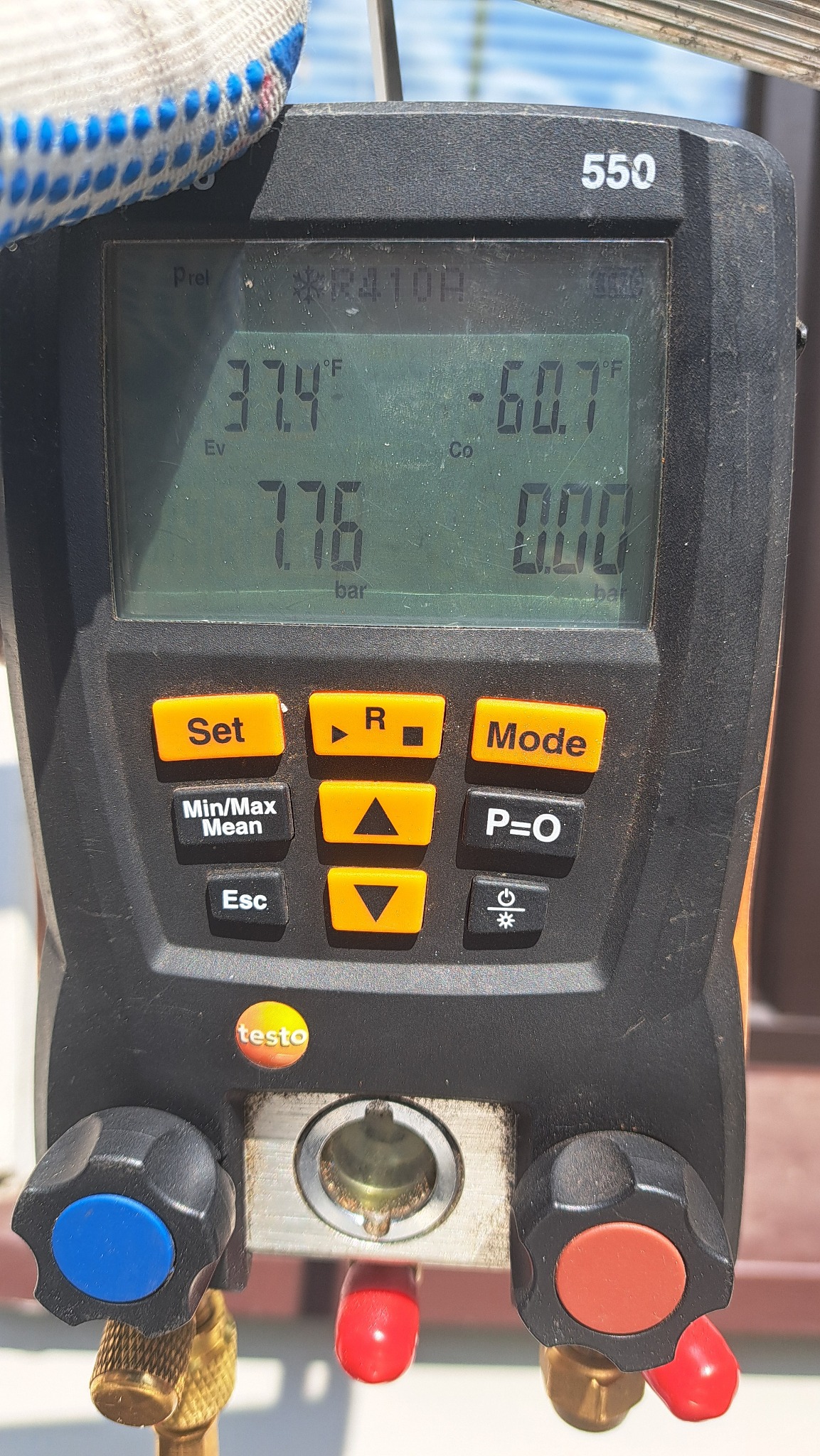 Cyfrowy manometr Testo 550, wskazujący wartości ciśnienia i temperatury, z widocznymi przyłączami i przyciskami funkcyjnymi, uchwycony w trakcie pomiaru, z fragmentem roboczej rękawicy w tle.