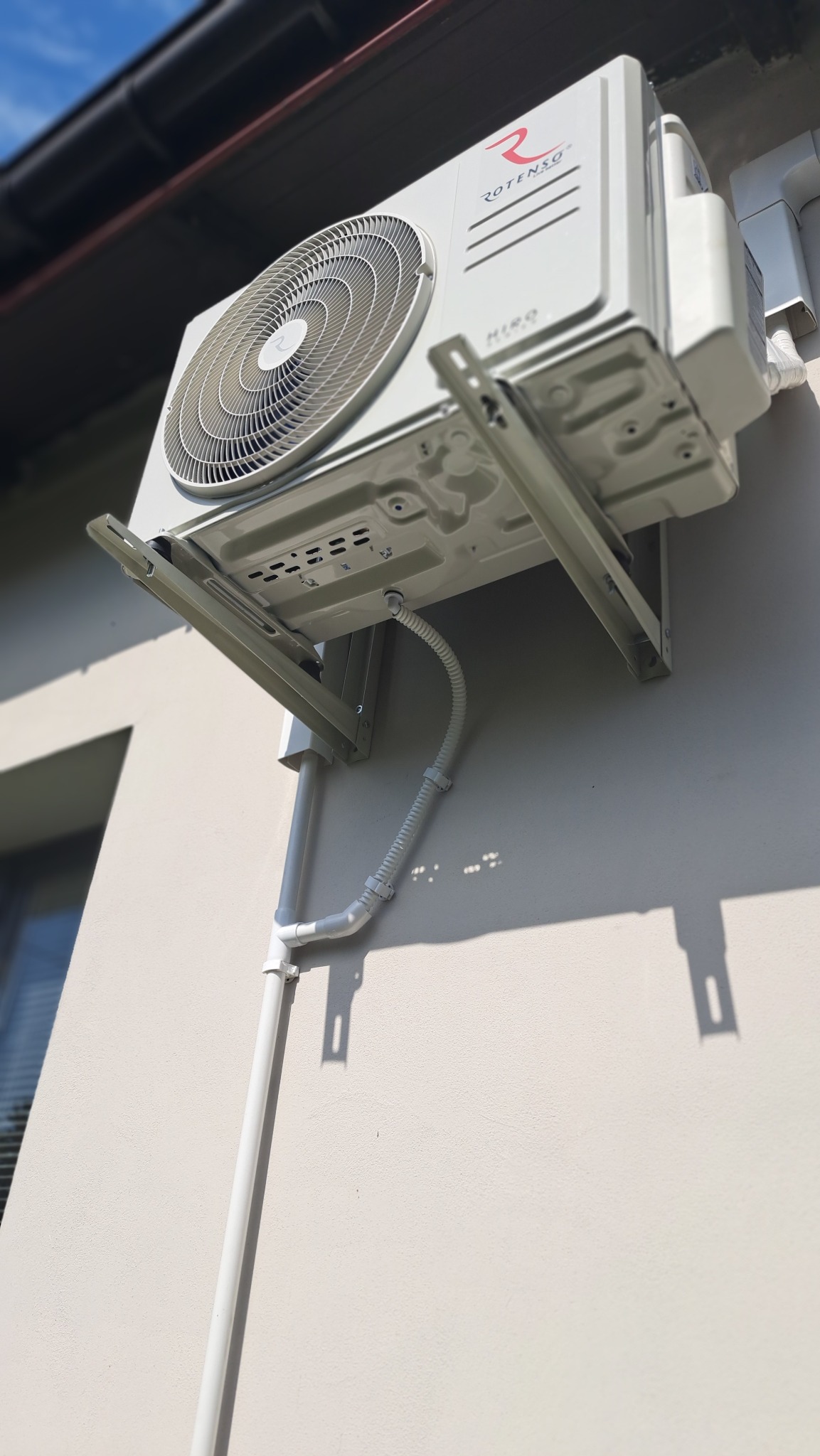 Biała jednostka zewnętrzna klimatyzacji Rotenso, zamontowana na ścianie budynku w Konstancin-Jeziorna, widoczna z dołu pod kątem, z widocznymi rurami odprowadzającymi.