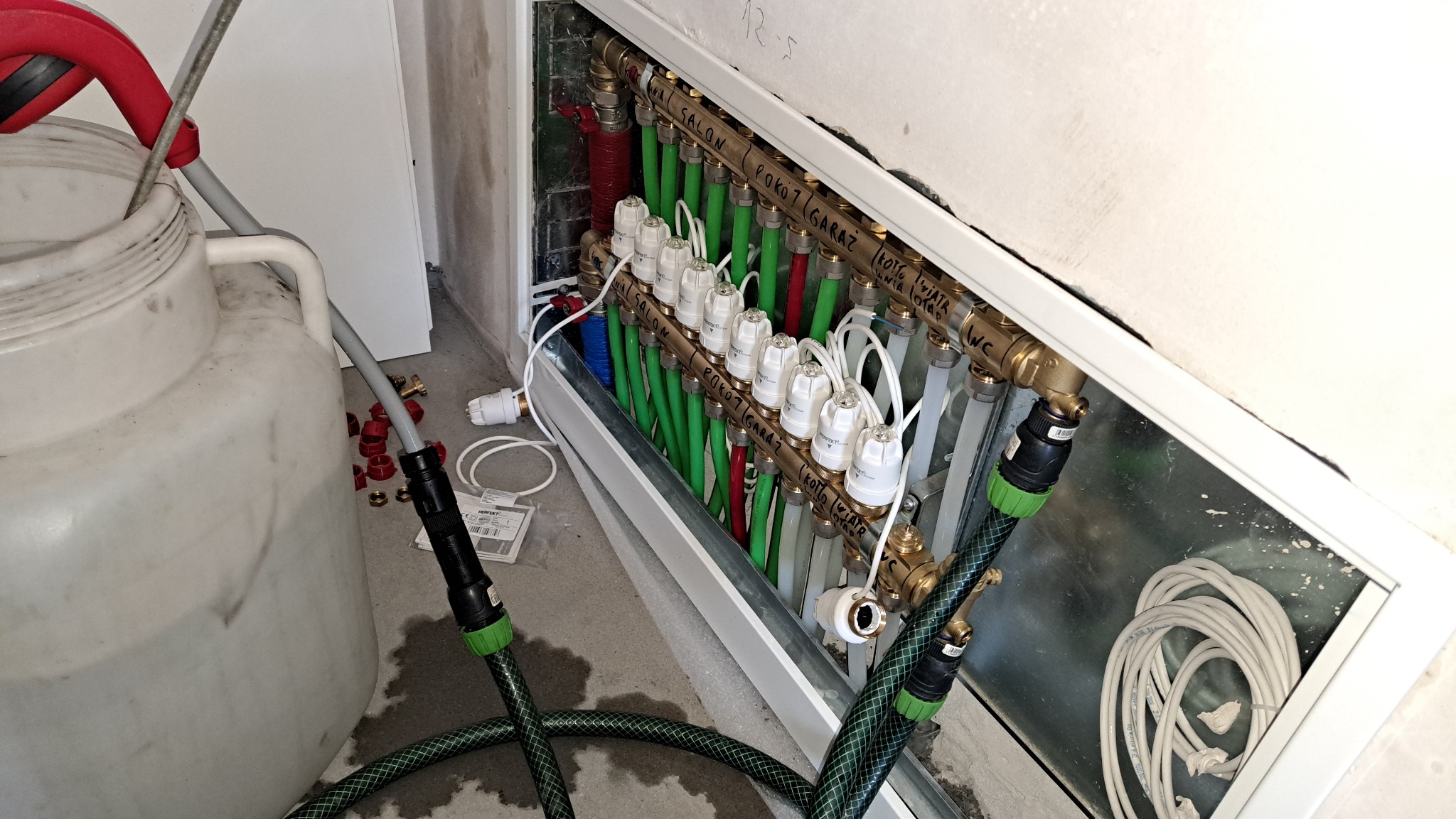 Rozdzielacz ogrzewania podłogowego z zielonymi rurami, podpisany nazwami pomieszczeń, podłączony do węży z zielonymi elementami, obok biały zbiornik i akcesoria instalacyjne.