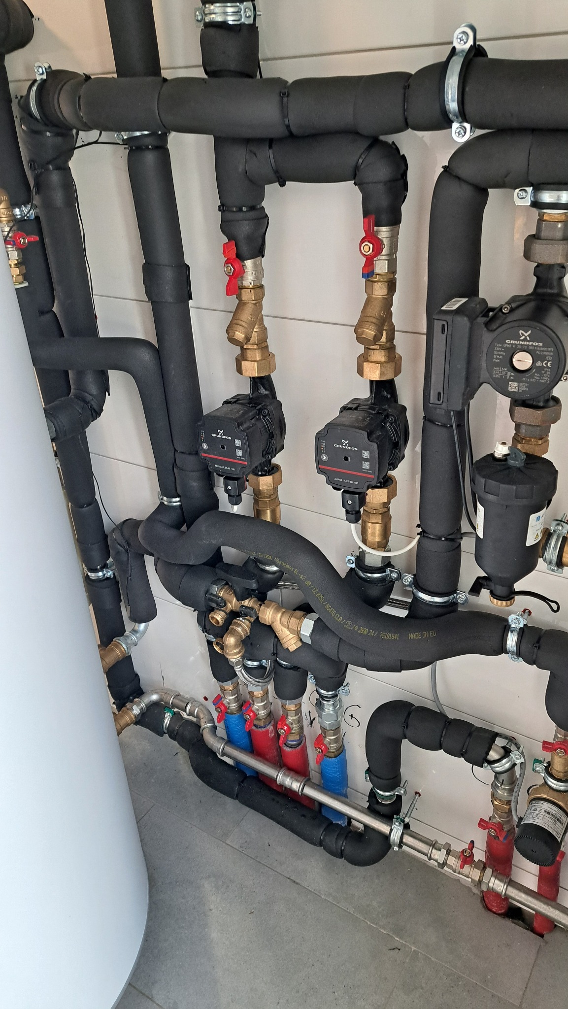 Instalacja grzewcza z dwoma pompami obiegowymi Grundfos Alpha L 25-40 180, rurami w izolacji termicznej, zaworami z czerwonymi pokrętłami i zbiornikiem akumulacyjnym.