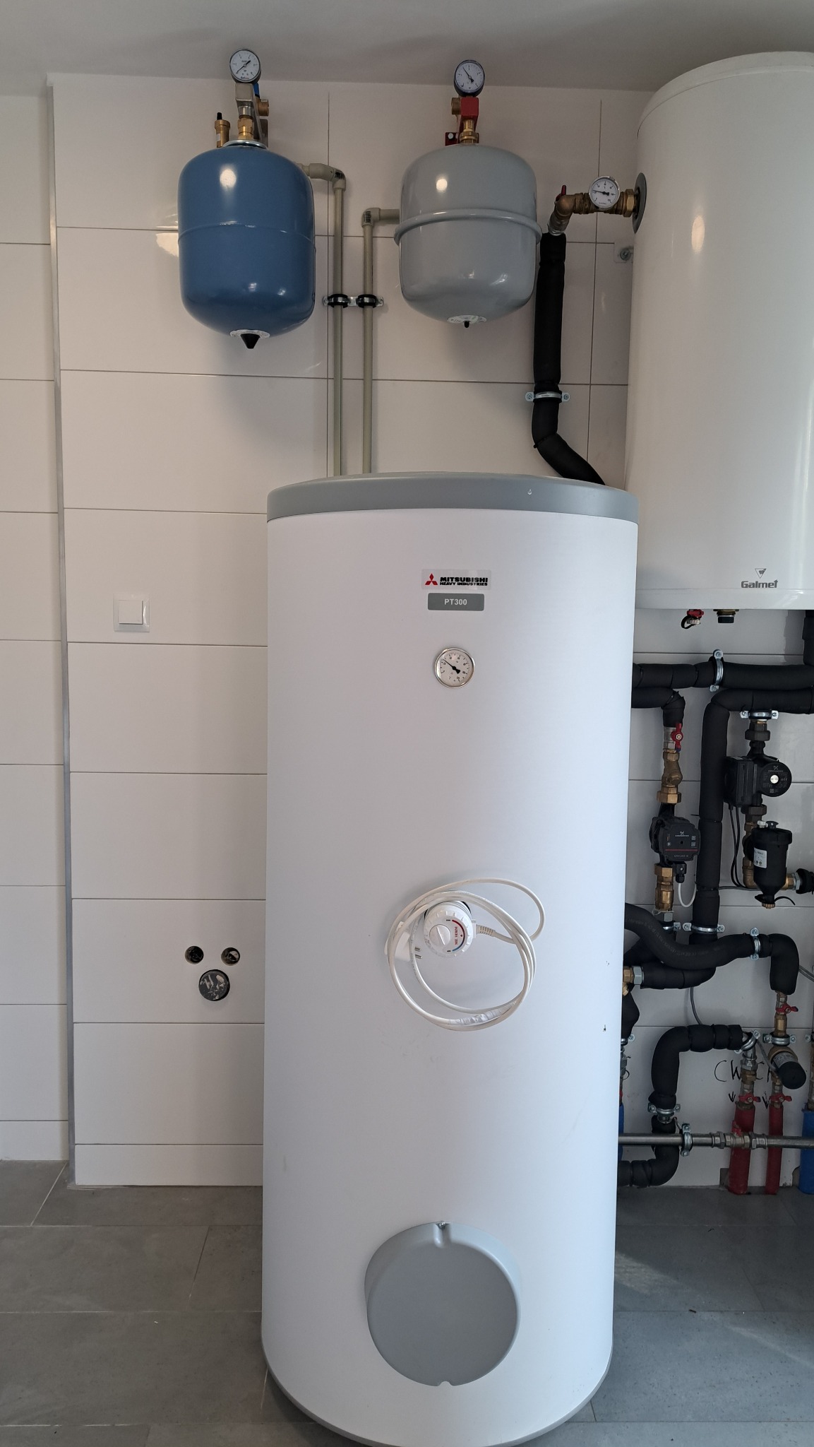 Biały zbiornik buforowy Mitsubishi PT300 z widocznym manometrem, termostatem i podłączeniami hydraulicznymi, zainstalowany w pomieszczeniu z białymi płytkami ściennymi i szarą podłogą.