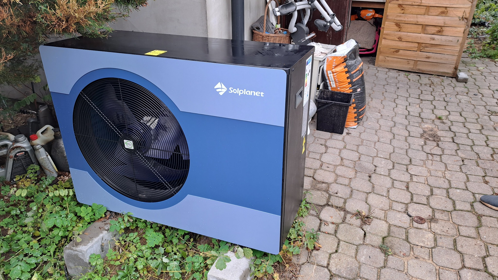 Zewnętrzna jednostka pompy ciepła Solplanet w odcieniach niebieskiego, umieszczona na betonowych bloczkach w otoczeniu zieleni, z widocznymi kanistrami oleju z lewej strony i workiem na ziemię w tle.