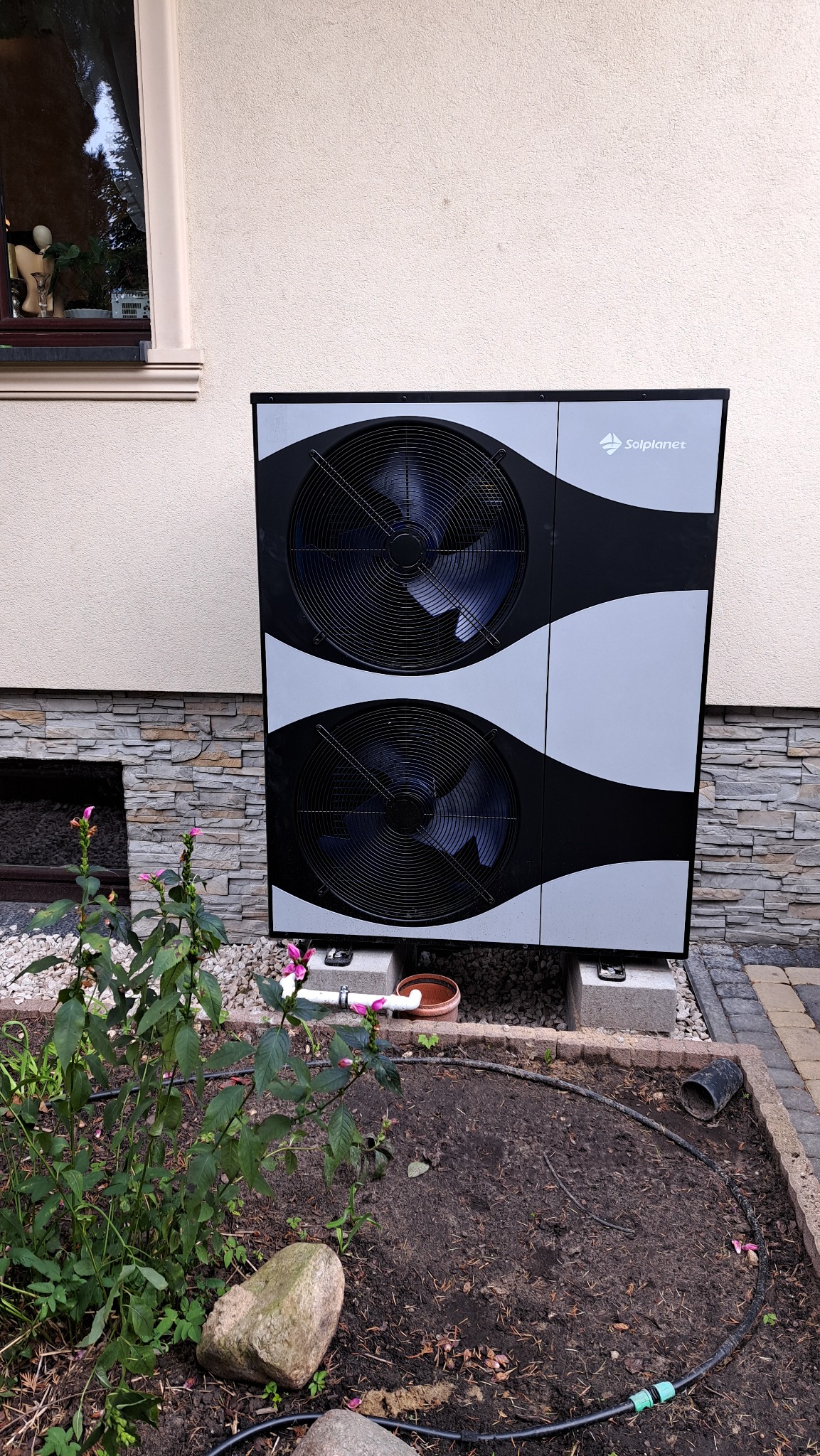 Zewnętrzna jednostka pompy ciepła Solplanet ustawiona na betonowych blokach przy ścianie domu z kamienną okładziną, obok rabata z roślinami i kamieniami.