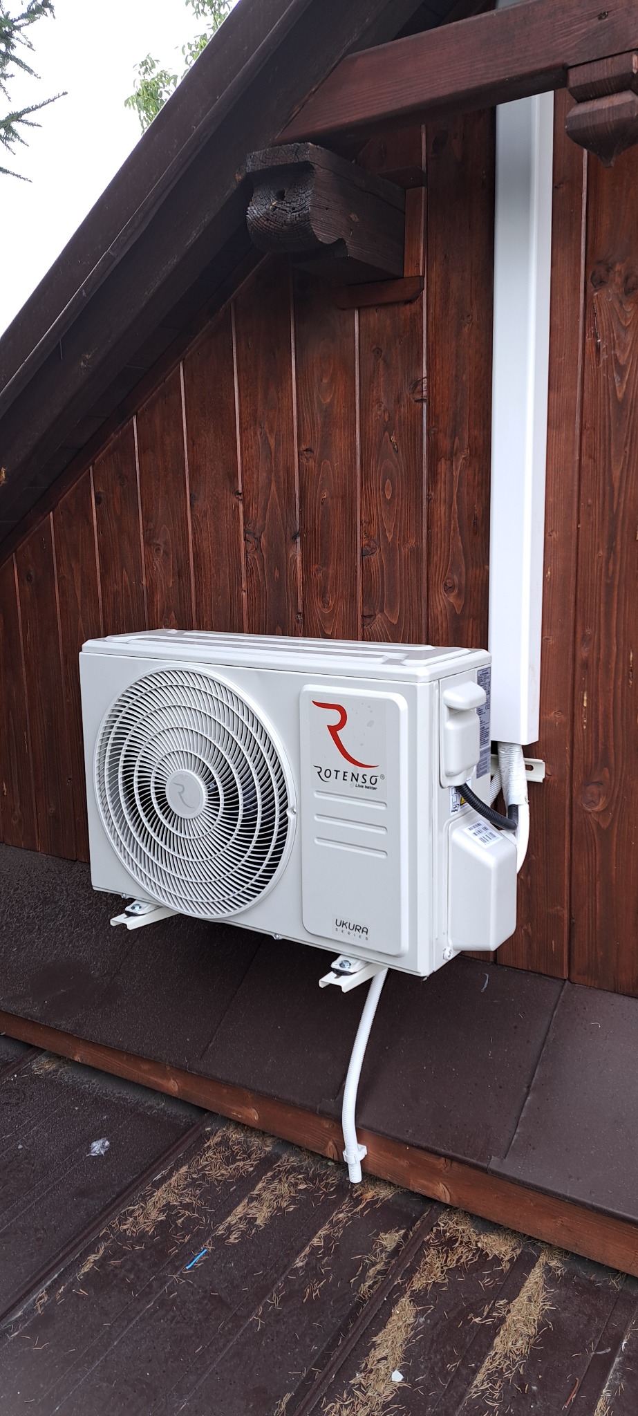 Zewnętrzna jednostka klimatyzacji Rotenso Ukura zamontowana na drewnianej ścianie budynku z widocznymi elementami instalacji odprowadzającej skropliny i osłoną przewodów.