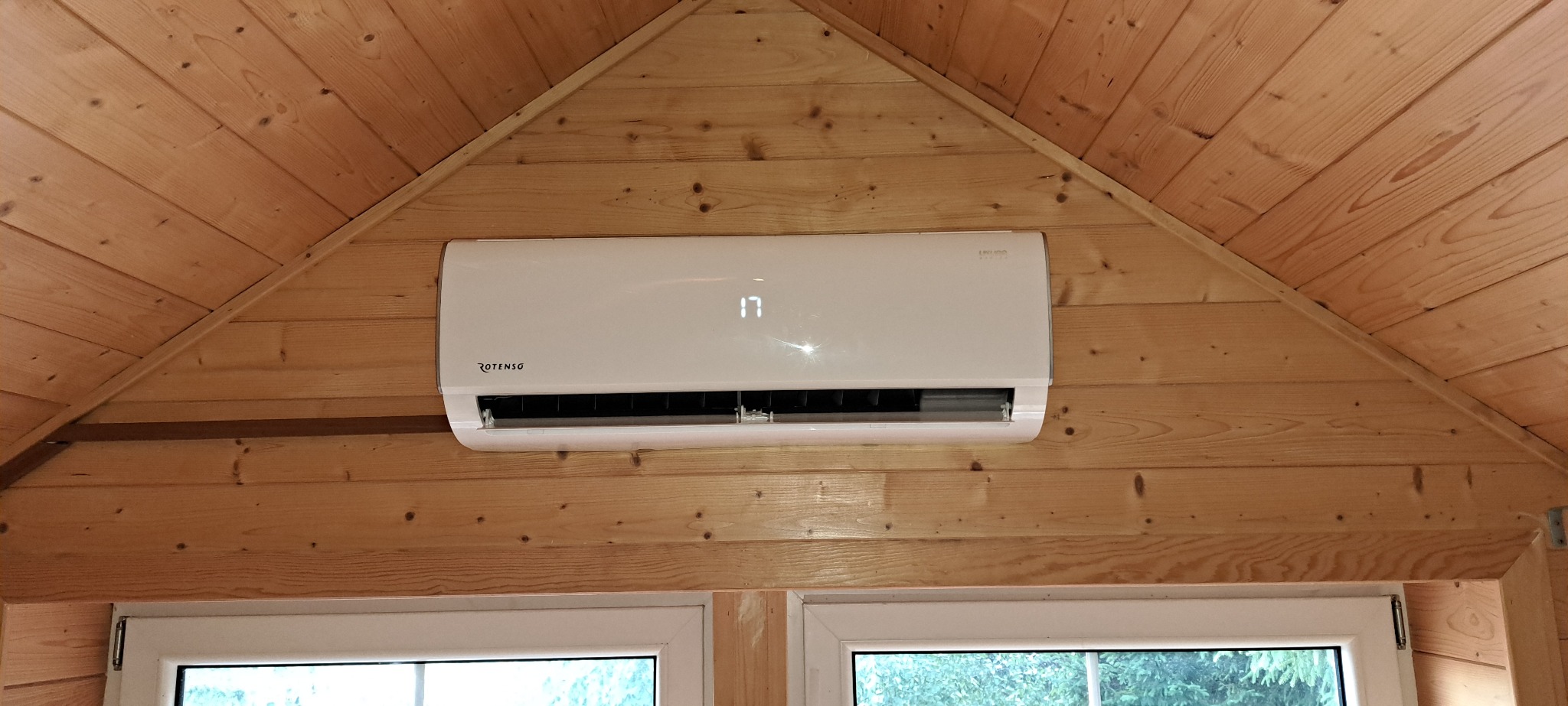 Biały klimatyzator marki Rotenso zamontowany na drewnianej ścianie w kształcie trójkąta, widoczne okno z zielenią na zewnątrz.
