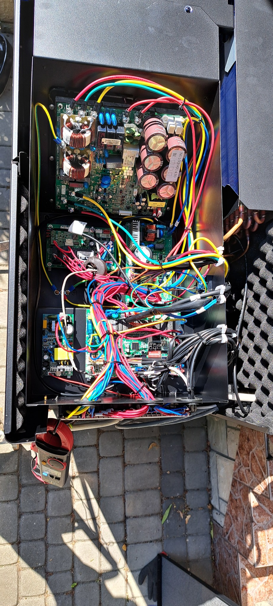 Wnętrze obudowy urządzenia z widocznymi płytkami drukowanymi, kondensatorami i wiązkami kolorowych przewodów, z multimetrem leżącym obok na brukowanej powierzchni.