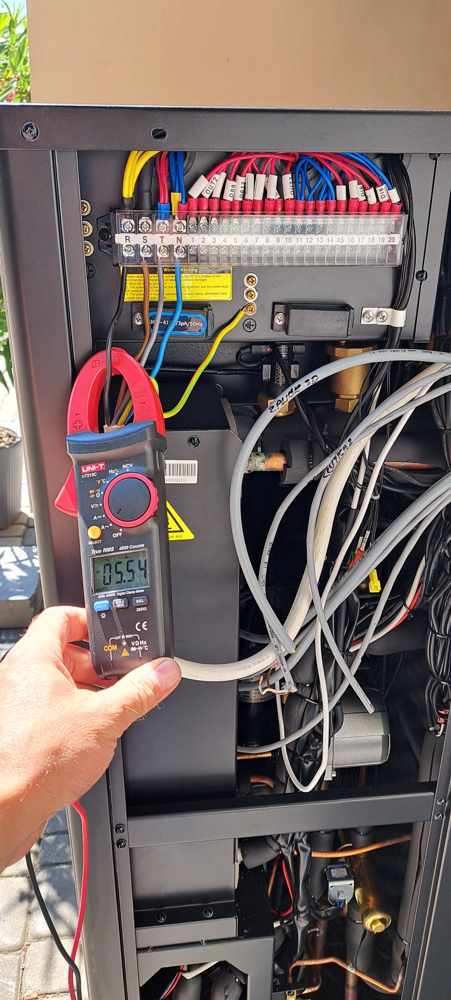 Pomiar prądu roboczego pompy ciepła za pomocą miernika cęgowego UNI-T UT213C, widoczne okablowanie i przyłącza elektryczne wewnątrz obudowy.